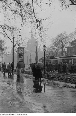 Menschen mit Regenschirmen warten an der Straßenbahnhaltestelle auf dem Martin-Luther-Ring, im Hintergrund das Reichsgericht