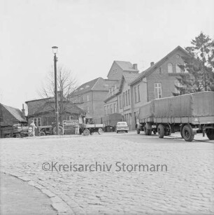 Klosterstraße, Kreuzung nach links Matthias-Claudius-Straße, nach hinten Ahrensböker Straße: Kopfsteinpflaster, Verkehrsinsel mit Straßenlaterne, Wegweiser nach Hamburg, Poller: zwei Lastwagen mit Anhängern, Pkw, Personen: Bildmitte hinten Claudius-Mühle, rechts Verwaltungsgebäude der Claudius-Mühle, Baum: links Kocksche Schmiede, Baum, Stuhlwagen, Traktor mit Anhänger