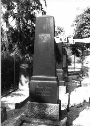 Grabstein 54 (Index: 99793123) Material: schwarzer Granit Zustand: gut Maße HxBxT: 191x38x21 cm Beschreibung: Obelisk, getreppter Sockel, Grabeinfassung: Grabstein 54