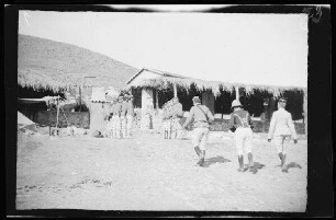 Nador (Marokko): 3 Offiziere in hellgestreiften Uniformhosen und unterschiedlichen Uniformjacken und Kopfbedeckungen "auf dem Weg zum Kommandeur" und spalierstehende Soldaten [des spanischen 8. Kavallerie-Regiments "Lusitania"] vor den Stallungen der 3. und 4. Eskadron (Rückansicht)