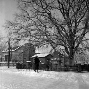 Lindenstraße: Ehrenmal mit Einfriedung und Baum: davor Frau mit Kind auf dem Arm: im Hintergrund Bauernhäuser: Winter