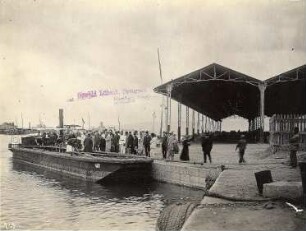 Ägypten. Touristen nach dem Anlegen in einem Hafen (am Nil oder Alexandria) mit offenen Lagerhallen für Waren, im Hintergrund Begleitboote der Hapag