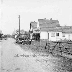 Ortseingang: Straßenschild und neue Wohnhäuser: links Stapel mit Baumstücken: dahinter Strommast: hinten links Pkw