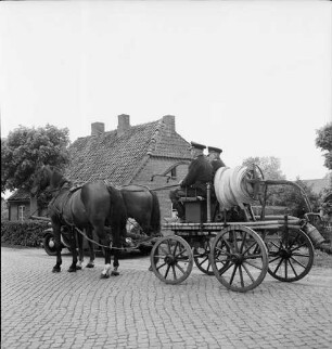 Freiwillige Feuerwehr Stubbendorf: Handdruckspritze von 1907 mit Pferdegespann: darauf zwei Feuerwehrmänner in Uniform: vorn Kopfsteinpflaster: dahinter Pkw: hinten Häuser und Bäume