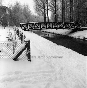 Beste: hinten Holzbrücke für Fußgänger: rechts Kurpark mit Bäumen: links eingezäunter Garten: dahinter Gebäude hinter Baumreihe: Winter