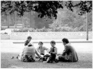 Eltern sitzen mit ihren Kindern auf einer Grünfläche (Altersgruppe 14-17)