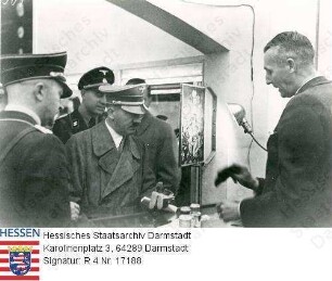 Hitler, Adolf (1889-1945) / Porträt in Staubmantel bei der Besichtigung einer Plexiglas/Acrylharz-Ausstellung in Mailand (Italien), Gruppenaufnahme, Halbfigur vor Glasvitrine