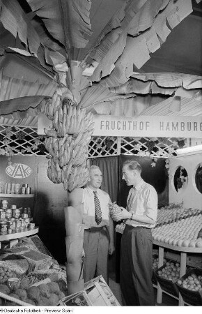 Zwei Männer umgeben von exotischem Obst auf der Leipziger Herbstmesse 1954
