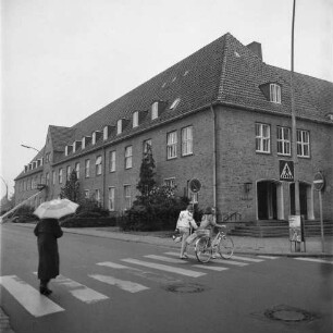 Altes Finanzamt [später Gebäude D]: Außenansicht: Blick von der Mommsenstraße: rechts Abzweigung Bangertstraße: vorn links Frau mit Regenschirm auf Fußgängerüberweg, davor zwei Mädchen mit Fahrrad