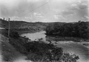 Flusslauf : Ein Fluss in Brasilien. Rechts Uferwald, links flaches mit Gras und Büschen bewachsenes Ufer. Im Hintergrund Hügel und links eine Straße.