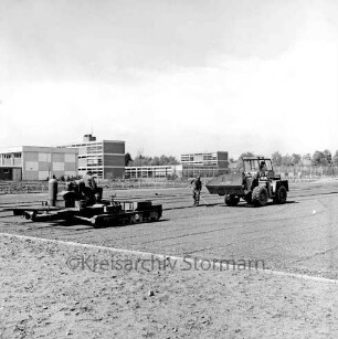 Sporteinrichtungen: Sportanlage am Kortenkamp: Ausbau: Hauptspielfeld: Auftragen des Hartbelages: Arbeiter mit Spezialfahrzeug und Bagger: hinten Emil-von-Behring-Gymnasium