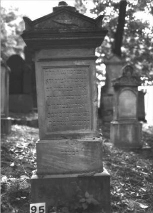 Grabstein 95 (Index: 99898135) Material: Sandstein Zustand: gut erhalten, Sockel etwas angewittert Maße HxBxT: 68x62x22 cm Beschreibung: aufgesockelte Stele mit Giebelaufsatz, im Giebelfeld Lorbeerkranz mit Schleife, unterer Sockel felsartig gearbeitet Symbolik: Lorbeerkranz: Grabstein 95