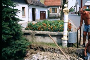 stillgelegter, mit Blumen bepflanzter Dorfbrunnen; Schachtarbeiten im Umfeld