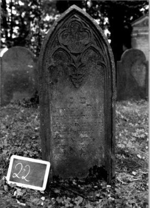 Grabstein 22 (Index: 100111048) Material: Sandstein Zustand: verwittert mit Textverlust Maße HxBxT: 110x46x14 cm Beschreibung: Stele, Spitzbogenabschluß, Schriftfeld eingetieft Symbolik: Maßwerk, Blatt: Grabstein 22