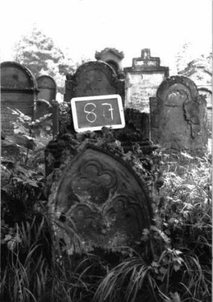 Grabstein 87 (Index: 100103556) Material: Sandstein Zustand: Stele in zwei Teile gebrochen, verwittert, vollständiger Textverlust Maße HxBxT: 163x56x19 cm Beschreibung: aufgesockelte Stele mit Spitzbogenabschluss, im Bogen Maßwerk, Schriftfeld eingetieft, in der Mitte durchgebrochen Symbolik: Maßwerk Besonderheit: ergänzt nach Gräberbuch: Grabstein 87