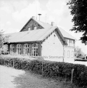 Volksschule: Außenansicht der Gebäude: vorn Hecke und Zaun: rechts oben Baumäste