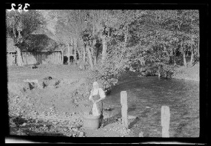 Domnetal (=Tal des Râul Doamnei, Fluss in Rumänien): Rumänin an einem Waschtrog neben einem Fluss, im Hintergrund ein Holzschuppen