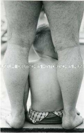Rückenansicht eines sitzenden Kindes zwischen den nackten Beinen eines stehenden Mannes