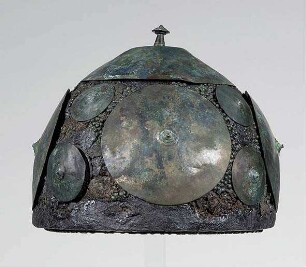 Helm aus Stücken wieder zusammengesetzt. Flechtwerk besetzt mit kleinen Bronzescheiben. Die Zwischenräume mit knopfartigen Stiften ausgefüllt (sog. Schüsselhelm)