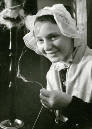 Mädchen aus Billigheim, lächelnd in Volkstracht, mit Haube gekleidet. Vor Spinnrad, einen Faden haltend. Berlin Pfalzausstellung. Portrait