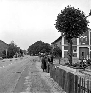 Alte Dorfstraße: vorn rechts Zaun: daran zwei Frauen: hinten Bauernhäuser und Grünanlage