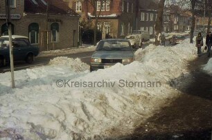 Königstraße: vorn parkender Pkw zwischen Schneehaufen: dahinter weitere Pkws: rechts zwei Personen: hinten Gaststätte und weitere Wohn- und Geschäftshäuser: hinten rechts vor Raiffeisenbank Kreuzung mit Hamburger Straße, Schützenstraße, Hagenstraße: Winter