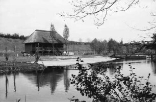 Trave: hinten links Sportlerhaus: davor Sandstrand: links Steg: hinten rechts Holzbrücke und Boot am Ufer: dahinter Bäume, Sportplatz und Hang am Trave-Stadion: vorn rechts Baumäste