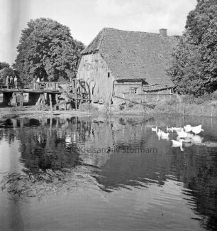 Grander Mühle: verfallenes Mühlengebäude und beschädigtes Mühlrad: links Holzbrücke: darauf Personen: vorn Mühlenteich: darauf rechts Gänse: hinten Bäume