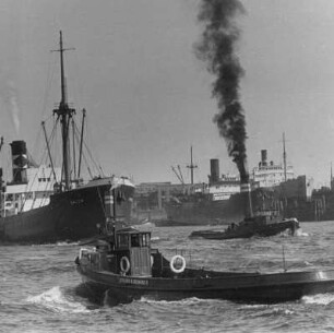 Hamburg. Im Hafen ist wieder emsige Betriebsamkeit. Schlepper bugsieren ein Frachtschiff aus dem Hafenbecken. Andere Schiffe liegen an den Kai´s. Im Vordergrund eine Fähre. Aufgenommen 1952