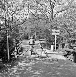 Lottbek: An der Lottbek: provisorisch ausgebesserte Holzbrücke über die Lottbek Richtung Hamburg: darauf Junge mit Fahrrad: links Straßenschild: rechts Ortsschild von Hamburg: dahinter Büsche und Bäume: hinten rechts Pkw