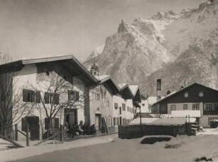 Oberbayern. Mittenwald im Winter. Verschneite Straße. Eine Gruppe von Menscehn sitzt vor einem Haus (eventuell Werkstatt). Im Hintergrund die Karwendelgebirgsgruppe