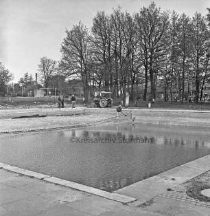 Freibad: vorn Schwimmbecken: dahinter Arbeiter mit Schaufel und Trecker bei Außenarbeiten: hinten links Sportplatz: hinten rechts Bäume und Häuser