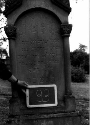 Grabstein 93 (Index: 100107259) Material: Sandstein Zustand: leicht verwittert, Aufsatz fehlt Maße HxBxT: 160x51x20 cm Beschreibung: ädikulaartige Stele mit Halbsäulen, getreppter Sockel, schräg eingezogener Rundbogenabschluß mit Verdachung, Schriftfeld mit Rundbogen Symbolik: Blattwerk, Rundbogenfries: Grabstein 93A