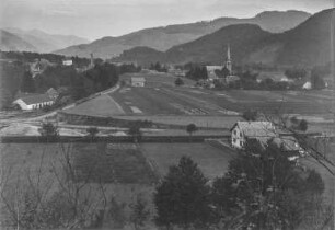 Dorf : Blick auf ein Dorf in einer Hochebene. Im Hintergrund bewaldete Berghänge und eine Kirche, im Vordergrund Felder und Weiden
