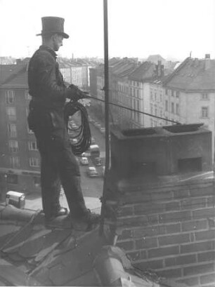 Schornsteinfeger beim Kehren eines Schornsteins, auf einem Dachgrat stehend