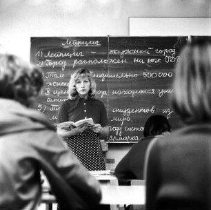 Russichlehrerin : Portrait einer Russischlehrerin, im Unterricht vor ihrer Klasse stehend mit aufgeschlagenem Lehrbuch in der Hand und Sätzen an der Tafel hinter ihr