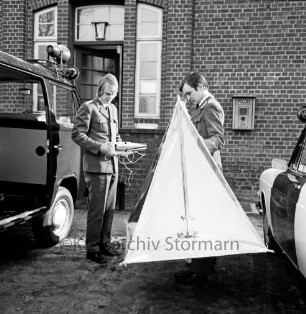 Polizeidienststelle: Alte Landstraße: Außenaufnahme, vor Eingang: Sicherstellen von Radiosonde des Deutschen Wetterdienstes Hamburg nach Absturz: zwei Polizisten mit Instrumentenkästchen und Flugkörper aus Aluminiumfolie: zu beiden Seiten Polizeifahrzeuge