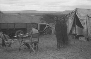 Zeltlager am Mount Hanang : Expeditionslager am Mount Hanang mit einem Expeditionsteilnehmer der an einem Tisch sitzt, mit dem Rücken zur Kamera und zwei Bewohnern die in die Kamera blicken