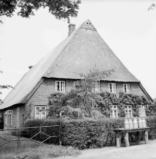 Bauernhaus mit Reetdach von Familie Orth: davor Vorgarten mit Hecke: vorn rechts Milchbock mit Milchkannen