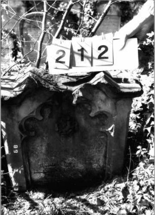 Grabstein 212 (Index: 99791541) Material: Sandstein Zustand: stark verwittert mit Textverlust Maße HxBxT: 60x60x23 cm Beschreibung: Stele, eingezogener doppelter giebelartiger Abschluß, Schriftfeld eingetieft Symbolik: Maßwerk, Blüte: Grabstein 212