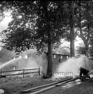 Gut Altfresenburg: Brand des Bullenstall: vorne ein geplatzter Wasserschlauch, rechts eine Feuerwehrmann, dahinter das brennende Gebäude