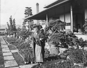 Yokohama. Botanischer Garten. Japanerin mit Kind auf dem Rücken im Bonsaigarten vor einem Gebäude