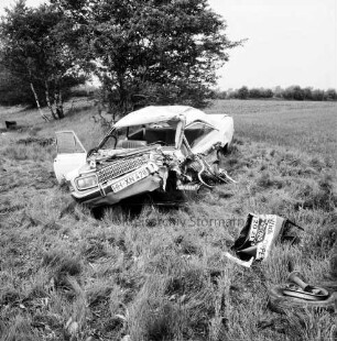 A 1: Verkehrsunfall: Zusammenstoß von Pkw und Lieferwagen: Wrack von Unfall-Pkw neben Straße, vorne Trümmerteil, Autorad: hinten Büsche, Bäume, Feld, Knick