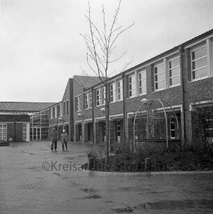 Bargteheide: Kreisgymnasium II [später Gymnasium Eckhorst]: Eckhorst: Außenaufnahme mit Schulhof: zwei Männer: vorne links Notausstieg aus Kellergeschoss, Tarnung als Brunnen