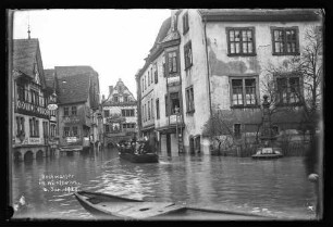 Hochwasser an der Ecke Rechte Tauberstraße/Lindenstraße mit Chorstift-Drogerie, Cafe Quenzer, Eisenhandlung A. Götzelmann u.a. Geschäften in Richtung Brückengasse, darauf Personen auf Schelch sowie Brunnen, darauf Schriftzug "Hochwasser in Wertheim 16. Jan. 1920"