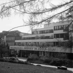 Krankenhäuser: Krankenhaus Reinbek St. Adolf-Stift: Hamburger Straße: Mittelbau mit Namenszug: davor Grünanlage mit Büschen, Bäumen sowie vorn rechts einer Plastik: links zwei Personen: dahinter Erweiterungsbau: rechts und oben Zweige eines Baumes