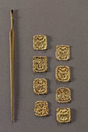 Spitz zulaufende Nadel, das obere Ende zu einem Löffelchen geformt, und acht quadratische Plättchen mit Kopf eines gehörnten Löwengreifen