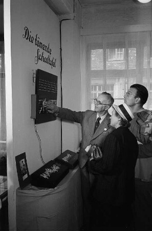 Drei Personen begutachten im Zentral-Messepalast auf der Leipziger Herbstmesse 1955 "Die tönende Schultafel" der Firma Doremi