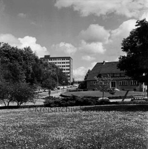 Kurpark: vorn Grünanlage mit Blumen, Hecken und Bäumen: dahinter Sülzberg: links Bangertstraße mit parkenden Pkws: dahinter Hochhaus [später Gebäude B] der Kreisverwaltung hinter Bäumen: rechts Gebäude mit Grünfläche