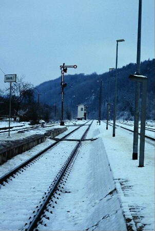 Bahnhof: verschneite Bahnsteige mit Schild, Signal und Gleis-Übergang